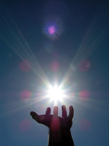 A silhouetted hand reaching toward a bright sun in a clear sky, with rays of light radiating outward.